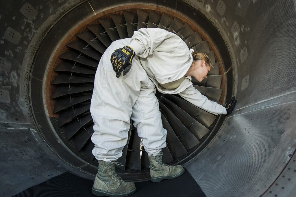 Jet propulsion technician in white coveralls bending inside a large turbofan engine intake to inspect the fan blades