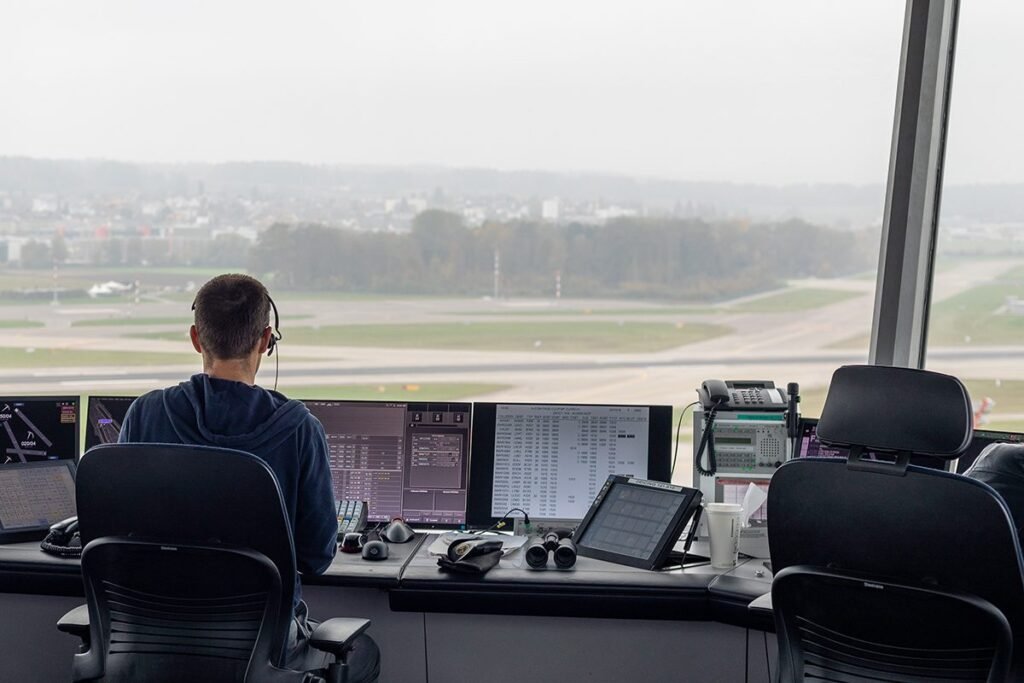Air traffic controller seen from behind at a console in an airport control tower, looking out over the runways