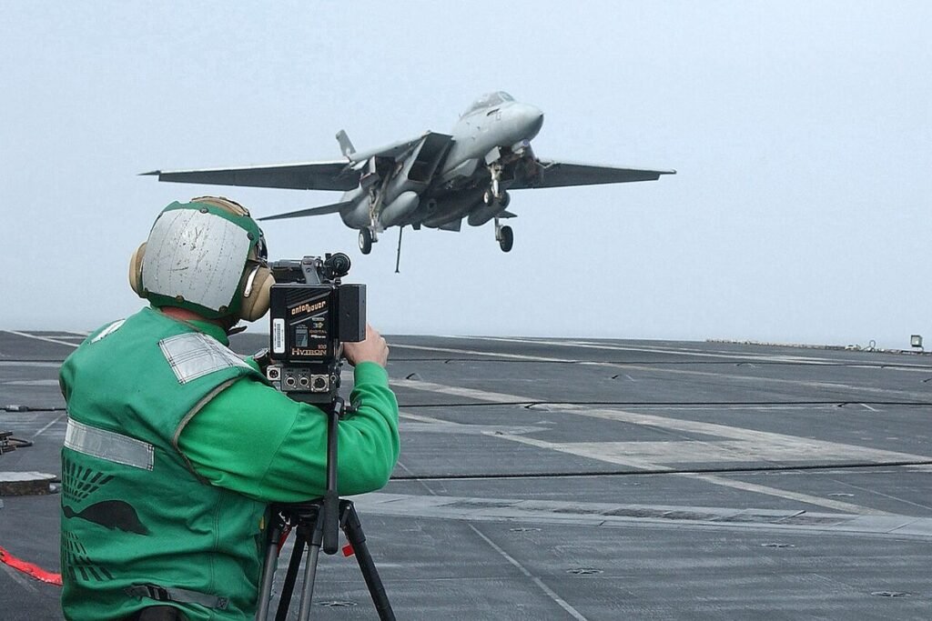 Navy videographer seen from behind filming an F-14 Tomcat as it launches from an aircraft carrier deck
