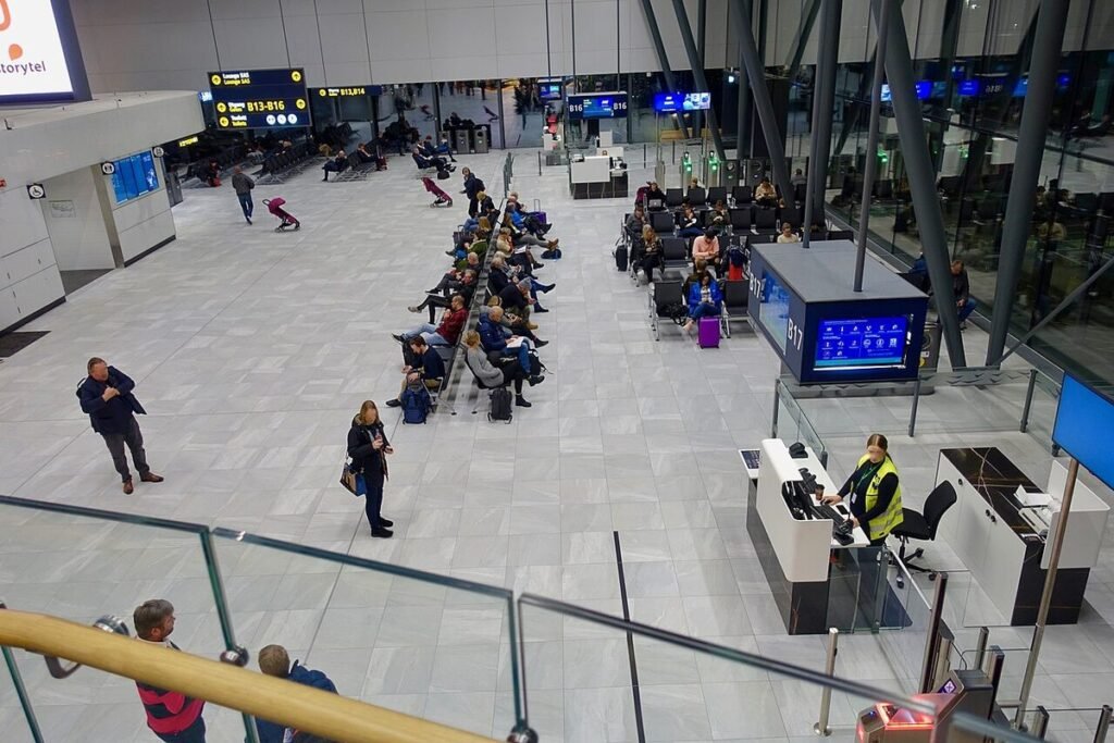 Passengers sitting and waiting in rows of seats at a departure gate in a modern airport terminal, seen from above