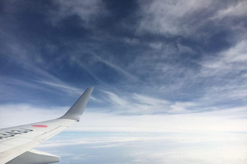 Airliner wing and winglet seen through a cabin window above a thin layer of clouds under a blue sky streaked with cirrus