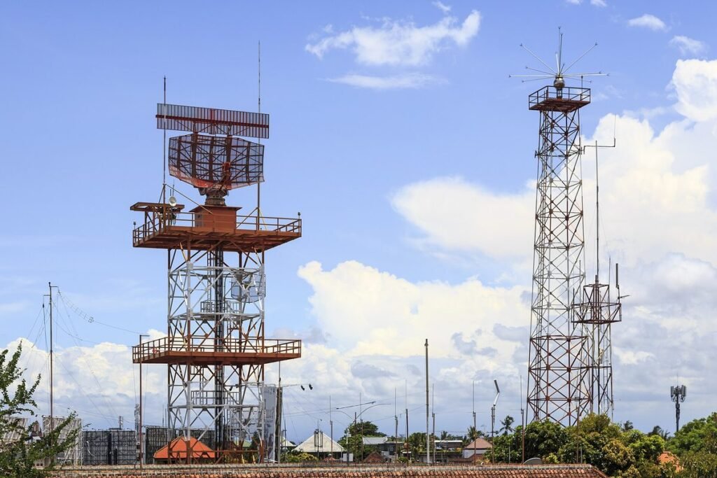 Airport surveillance radar antenna on a steel tower next to a navigation antenna mast under a blue sky