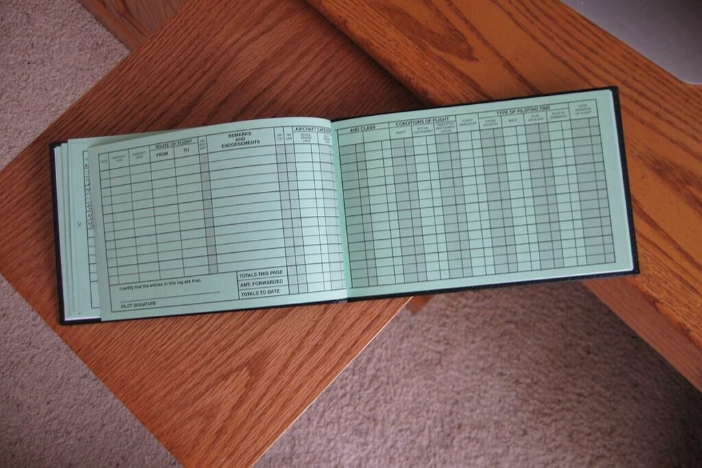 Open paper pilot logbook showing ruled columns for flight time entries, lying on a wooden table