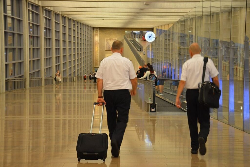 Two airline pilots in white uniform shirts seen from behind, walking down a long terminal corridor with a roller bag and flight bag