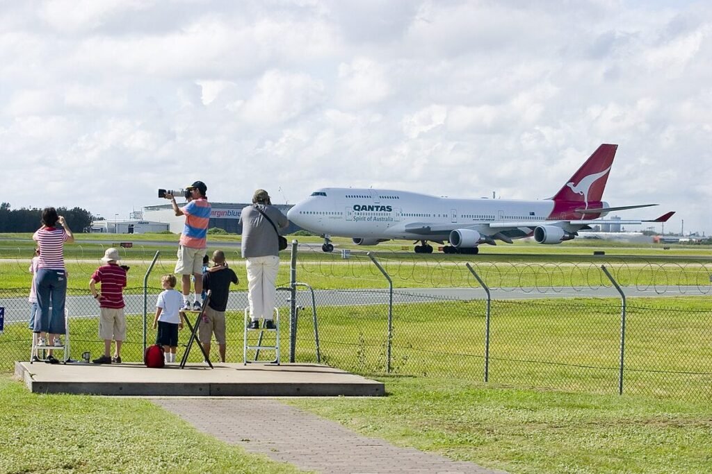 Aircraft spotters on a raised viewing platform, one with a long telephoto lens, photographing a Qantas Boeing 747 on the taxiway